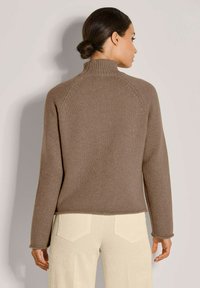 Brauner gestrickter Pullover mit hohem Kragen, gerippter Textur und Raglanärmeln. Kombiniert mit hellen Cordhosen.