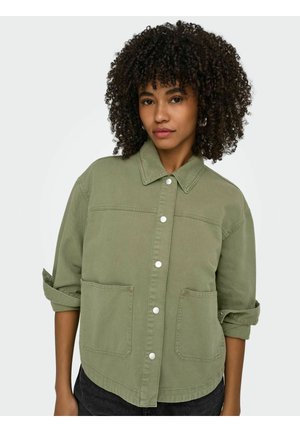 Femme aux cheveux bouclés portant une veste boutonnée vert olive avec les manches retroussées et de grandes poches frontales, posant devant un fond uni.