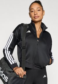 Črna jakna z zadrgo in belimi črtami na rokavih, izdelana iz gladkega blaga. Ima visok ovratnik in bočne žepke. Nosilna črna športna torba Adidas.