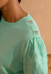 Piparmētru zaļš adīts džemperis ar viļņotu kakla izgriezumu un mežģīņu raksta pūkainām piedurknēm. Dekoratīvas zeltainas lentītes uz pleca piešķir detaļas.