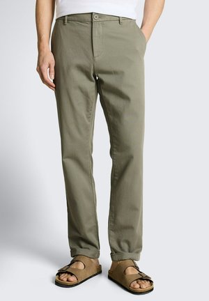 Homme portant un pantalon droit vert olive avec des ourlets retroussés et des sandales marron à bout ouvert avec boucle, debout avec une main dans la poche.