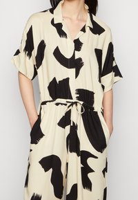 Mono de patrón negro y beige con mangas cortas, cuello, cordón en la cintura y dos bolsillos laterales. Textura de tela suave.