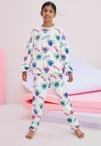 Valkoinen fleece-pyjama-asuste, jossa on värikkäitä piirroskuvioita Stitchistä. Setissä on pitkät hihat ja housut, joustavat hihansuut sekä pehmeä tekstuuri.