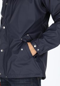 Marineblaue wasserfeste Jacke mit glatter Textur, Druckknöpfen und einer Seitentasche. Manschetten mit Druckknöpfen gesichert, entworfen für Funktionalität und Komfort.