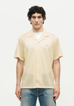 Νέος άνδρας με σγουρά σκούρα μαλλιά φοράει ένα μπεζ κοντομάνικο πουκάμισο πάνω από ένα λευκό μπλουζάκι, συνδυασμένο με μπλε τζιν, σε απλό φόντο.