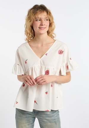Junge Frau mit lockigem blondem Haar, die eine weiße Bluse mit rosa Blumenmuster und hellblaue Jeans trägt, steht vor einem weißen Hintergrund.