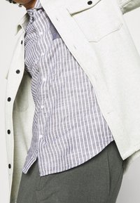 Ljusgrå knappad kappa med svarta knappar över en blå och vit randig skjorta. Grå byxor fulländar outfitet.