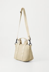 Beige taske lavet af tekstureret stof med to håndtag øverst og en lang justerbar rem. Har logo-detaljer på remmen og sort hardware.