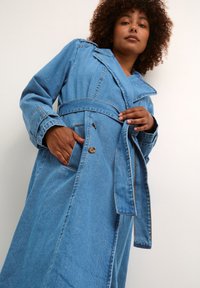 Kvinna med lockigt hår som bär en blå jeansjacka med skärp, ena handen i fickan och den andra justerar skärpet, mot en enfärgad vit bakgrund.