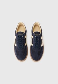 Adidași din suede bleumarin cu o dunga crem, design cu șiret, și talpă din cauciuc bej. Partea superioară texturată cu accente contrastante.