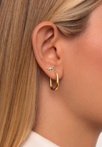 Goldene Creole kombiniert mit einem kleinen Libellen-Ohrstecker, der einen blau-grünen Körper und einen klaren Edelsteinakzent zeigt, was eine Mischung aus Texturen und Farben präsentiert.