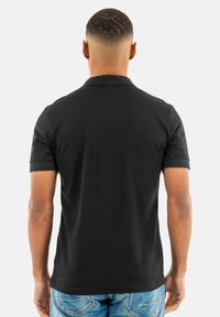 Polo negro de manga corta, con collar clásico y tejido de algodón suave. Llevado con jeans de color azul claro, visto desde atrás.