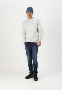 Felpa grigia con polsini a coste, cappellino blu, jeans in denim scuro e sneaker nere. Loghi distintivi sulla felpa e sul cappellino.