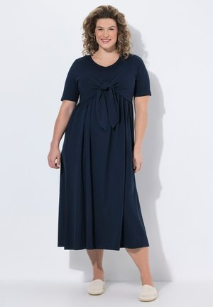 Vrouw met krullend haar die een marineblauwe midi-jurk draagt met een strikdetail aan de voorkant en witte slip-on schoenen, staand tegen een witte achtergrond.