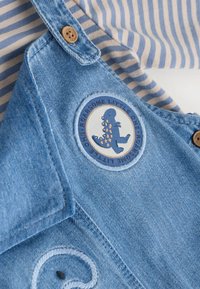 Prenda de denim con botones de madera y un parche de dinosaurio que dice "Increíble Pequeño" sobre un fondo de tela a rayas azul y beige.