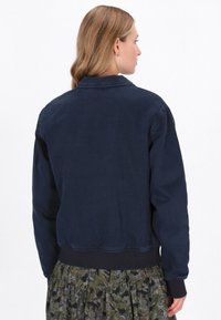 Tengerészkék farmerdzseki, gallérral, hosszú ujjal és bordázott alsó szegéllyel. Egyszerű dizájn, díszítések nélkül.