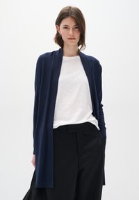 Marineblå cardigan i mykt stoff, åpen frontdesign med lange ermer, matchet med en hvit T-skjorte og svarte bukser. Enkel, flytende silhuett.