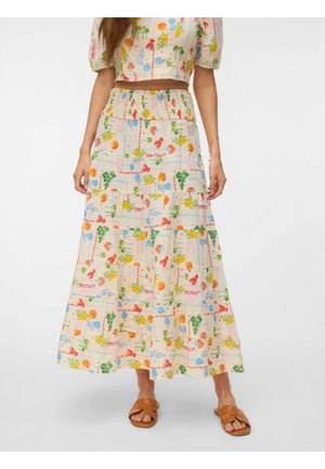 Jeune femme portant un crop top crème assorti et une longue jupe avec un imprimé tropical coloré, debout les mains sur les hanches, sandales plates couleur fauve.