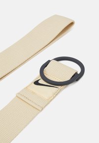 Beige neulottu kangasvyö, jossa on musta pyöreä solki ja beige etiketti, jossa on musta Nike-logo. Raskaasti teksturoitu pinta, kestävä materiaali.