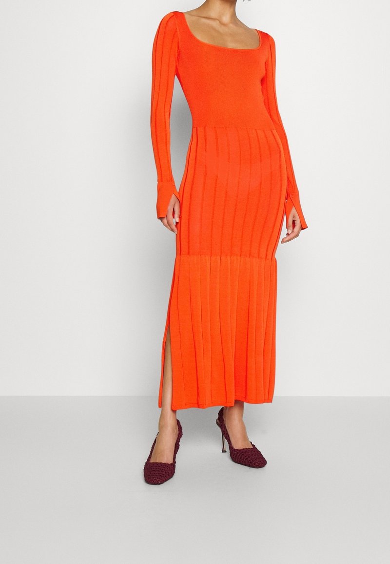 Femme portant une robe tricotée côtelée à manches longues de couleur orange vif avec des fentes sur les côtés et des chaussures à talons hauts bordeaux, debout devant un fond uni.