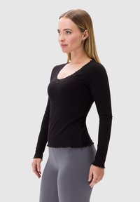 Czarny longsleeve z prążkowanej tkaniny i koronkowym dekoltem w kształcie V; ma falbaniasty dół i dopasowany krój, noszony z szarymi legginsami.