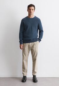 Blauer gerippter Pullover mit Rundhals, cremefarbene tapered Hose und schwarze Stiefel bis zum Knöchel. Vor einem schlichten weißen Hintergrund.