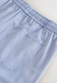 Gros plan sur un pantalon bleu clair montrant une ceinture élastique et une unique poche plaquée arrière avec des détails de couture visibles.