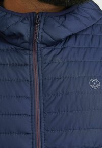 Námornícky pufový kabát s horizontálnym prešívaním, ktorý má predný zips, kapucňu a kruhové logo s nápisom "Jack & Jones" na hrudi.