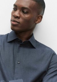 Marineblå, struktureret skjorte med knaplukket krave, der har et lille ternet mønster og en enkelt knap på frontknapstolpen.