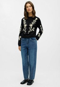 Pull noir avec broderie florale crème, associé à un jean bleu ample et des chaussures en cuir noires. Design simple, tissu de poids moyen.