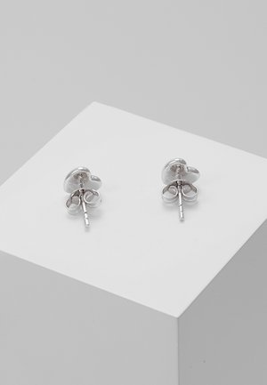 Petites boucles d'oreilles clous argent en forme de cœur avec attaches papillon, présentées sur un cube blanc sur un fond gris.