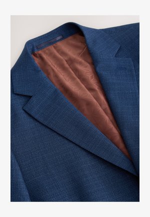 Mørkeblå blazer med tekstureret ydre, der har en brun inderside med geometrisk mønster. Bemærkelsesværdig peak lapel og skræddersyet form.