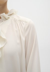 Krémová blůza s volánkovým límečkem, dlouhými rukávy a texturovaným, lehkým materiálem. Design zahrnuje jemné řasení na ramenech.