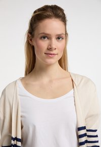 Cardigan beige con righe blu navy sulle maniche, indossato su una camicia bianca. Texture lavorata a maglia, design aperto sul davanti, silhouette aderente.