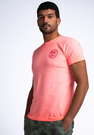 T-shirt di cotone rosa con scollo rotondo, maniche corte e stampa grafica di una palma sul petto sinistro. Indossato con pantaloni corti a fantasia mimetica.