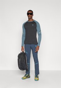 Maglietta nera a maniche lunghe con dettagli blu, abbinata a pantaloni navy. Il modello tiene uno zaino nero. Le scarpe sono gialle con dettagli blu.