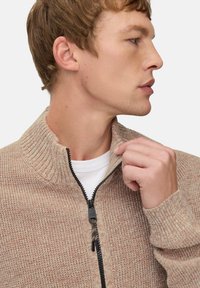 Beige gestrickter Pullover mit einem Halb-Reißverschlusskragen, gerippter Textur und schwarzem Reißverschluss, kombiniert mit einem weißen Hemd darunter.