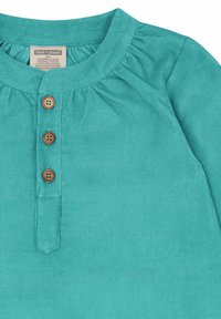 Top in cotone ribbed color teal con scollatura rotonda, dettagli arricciati e quattro bottoni in legno alla patta. Texture morbida.