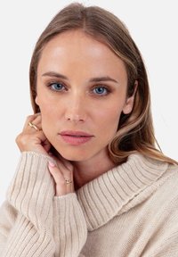 Femme aux yeux bleus portant un pull en tricot beige, reposant le menton sur des mains jointes ornées de bagues en or, fond neutre.