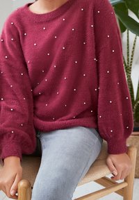 Burgunderfarbenes, weiches, flauschiges Sweatshirt mit Ballonärmeln, verziert mit verstreuten kleinen silbernen Perlen, getragen über hellgrauen Jeans.