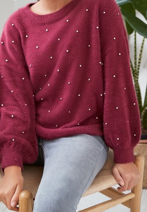 Burgundská, měkká, plyšová svetr s balónovými rukávy, zdobený rozptýlenými malými stříbrnými korálky, nošený přes světle šedé džíny.