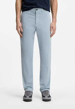 Uomo che indossa pantaloni azzurri a gamba dritta, camicia blu navy e sneakers grigie e blu navy, in piedi con le mani rilassate lungo i fianchi.