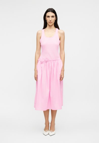 Woman with long dark hair wearing a sleeveless pink midi dress with floral detail at the waist and white pointed-toe heels, standing still.