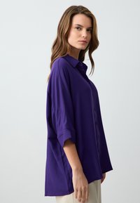 Chemise violette à boutons, en tissu lisse, dotée d un col, de manches retroussées et d une coupe ample. Design simple sans motifs visibles.