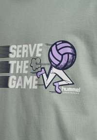 Ljusgrön bomullströja med ett grafiskt motiv av en springande figur med en lila volleyboll som huvud och texten "SERVE THE GAME."