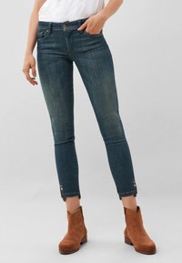 Jeans ajustados en denim azul oscuro con áreas deslavadas, diseño de cinco bolsillos, dobladillos acentuados con detalles de botones, combinados con botas de tobillo marrones.