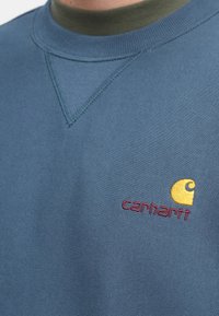 Modrá mikina z měkké látky, s kulatým výstřihem a žlutým vyšitým logem s červeným textem na hrudi.