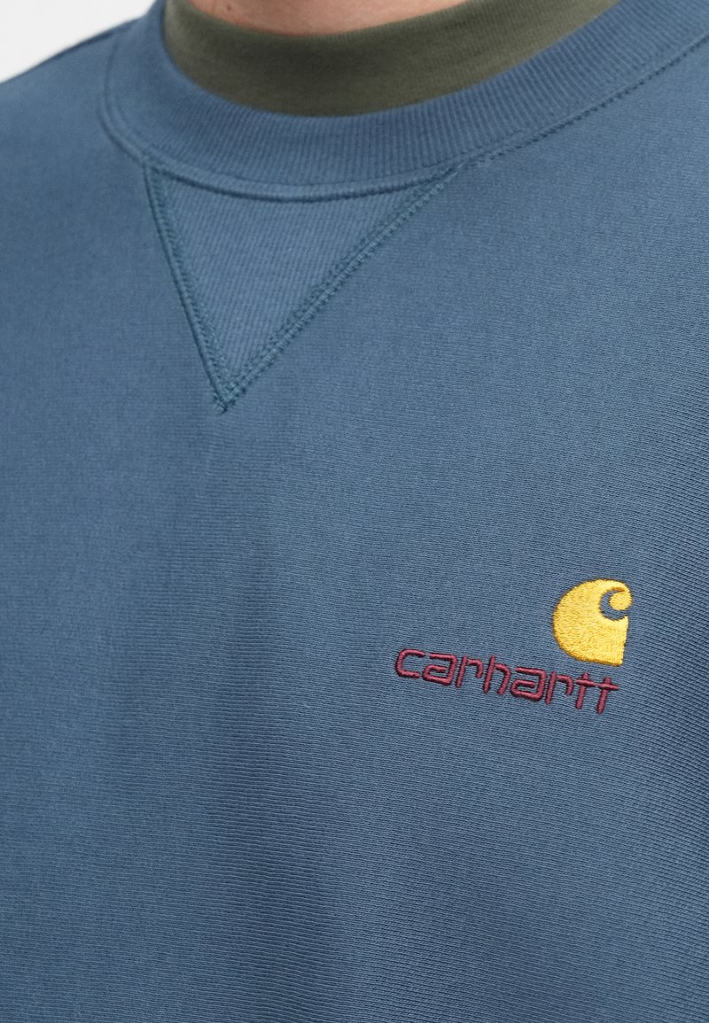 Blå sweatshirt lavet af blødt stof med rund halsudskæring og en gul broderet logo med rød tekst på brystet.