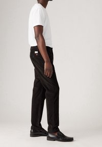 Svarta kordflortbyxor med rak passform, kombinerade med en åtsittande vit t-shirt och svarta läder loafers; inkluderar en liten röd etikett på bakfickan.