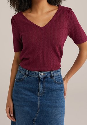 Vrouw draagt een bordeauxrode, getextureerde V-hals top met korte mouwen, ingestopt in een hooggesneden blauwe denim rok met voorzakken en knoopsluiting.